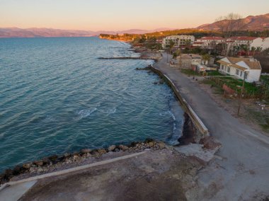 Yumuşak dalgalı sakin kıyı manzarası, renkli gün batımı ve kıyıda dizili binalar..