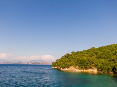 Berrak bir gökyüzü altında yemyeşil ve sakin mavi sularla sakin bir sahil manzarası..