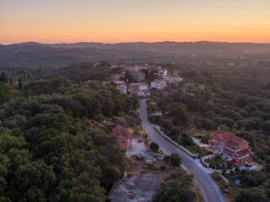 Yuvarlanan tepelerin ve yemyeşil yeşilliklerin arasına kurulmuş huzurlu bir köyün hava günbatımı manzarası.