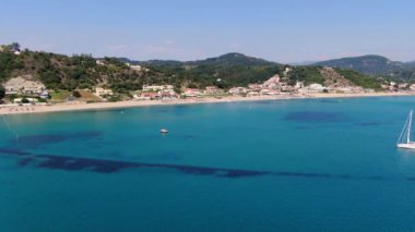 Kumlu bir sahil ve tatil köyü yakınında yelkenli ve rüzgar sörfçüleriyle turkuaz bir okyanusun havadan görünüşü. Korfu, Yunanca