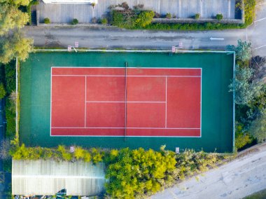 Ağaçlar ve binalarla çevrili canlı kırmızı ve yeşil tenis kortunun havadan görünüşü.