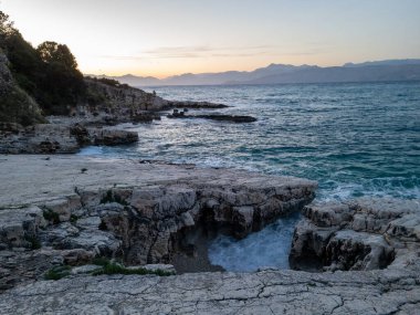 Kıyı günbatımı manzarası, kayalık kıyı sakin denizle buluşuyor. Dalgalar yavaşça engebeli kıyı şeridine çarpar ve sakin bir atmosfer yaratır..