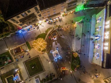 Şehir meydanının gece manzarası. Tatil ışıkları ve süslemelerle güzelce aydınlatılmış. İnsanlar şenlikli atmosferde geziniyor..