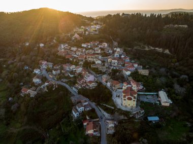 Tepe kenarındaki bir köyde gün batımı, kıyı şeridine doğru çağlayan evler. Altın saat ışığı sahneyi aydınlatıyor.