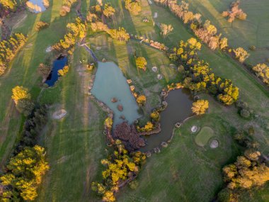 Göletli ve ağaçlı bir golf sahasının havadan görünüşü. Altın saat güneş ışığı yeşilliklerin üzerine gölge düşürüyor.
