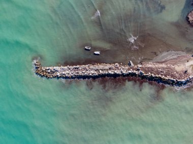 Turkuaz denize uzanan kayalık bir dalgakıranın havadan görünüşü. Kıyıda iki küçük tekne görünüyor..