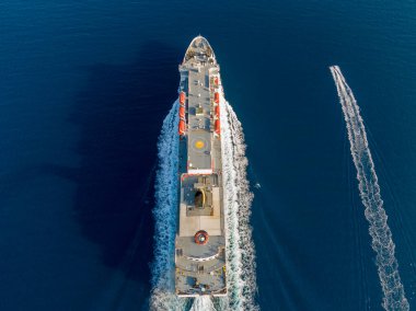 Büyük bir kargo gemisi açık okyanusta seyrediliyor, yukarıdan. Daha küçük bir gemi arkasında iz bırakıyor..