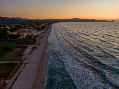Bir sahil kasabası ve sahil üzerinde gün batımı. Dalgalar kıyıya vuruyor, huzurlu bir manzara yaratıyor..
