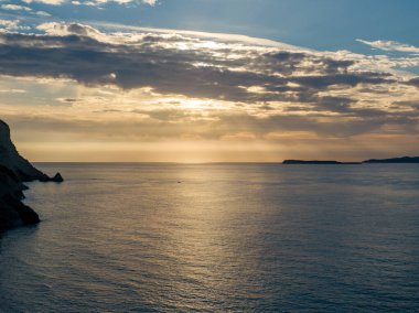 Okyanusun üzerinde sakin gün batımı, sakin sulara yansıyan altın ışık. Karanlık uçurumlar ve gökyüzüne karşı uzak bir ada silueti.