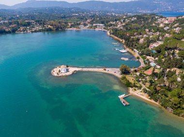 Küçük ada ve kilisesiyle kıyı manzarası, berrak turkuaz su ve arka planda yemyeşil tepeler..