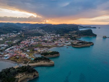 Gün batımında sahil kasabası, hava manzaralı. Turkuaz sular, dramatik gökyüzü ve kayalık kıyı şeridi çarpıcı bir manzara yaratır..