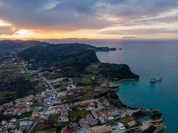 Yeşil tepeler ve okyanus arasında yuva yapmış bir kasabanın sahil gün batımı manzarası.