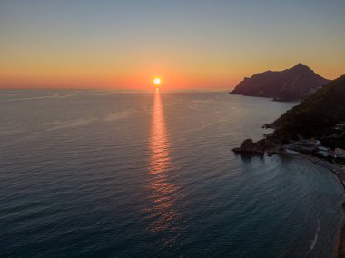 Okyanusun üzerinde gün batımı, suya yansıyan altın saat ışığı. Arka planda kıyı sıradağları.