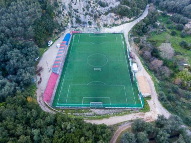 Yeşil bir arazide yuva yapmış bir futbol sahasının havadan görünüşü. Etrafı ağaçlarla ve toprak yollarla çevrili, sakin bir spor manzarası sunuyor..