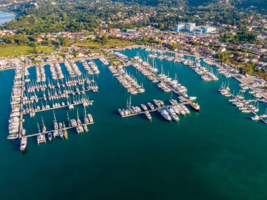 Yat ve yelkenlilerle dolu bir marinanın havadan görünüşü, düzenli bir şekilde kenetlenmiş. Arka planda bir kıyı kasabası ve yeşil tepeler görülüyor..