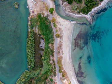 Sakin bir sahil manzarasının havadan görüntüsü. Turkuaz su, yemyeşil ve yollarla çevrili kumlu bir plajla buluşur..