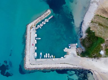 Sakin bir limanın havadan görünüşü, kayıklar düzgün bir şekilde bir taş iskeleye yanaşmış. Turkuaz su, huzurlu marinayı çevrelemektedir..