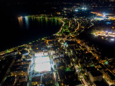 Kıyı şehrinin gece görüşü, aydınlık sokaklar ve karanlık bir su kütlesinin etrafındaki binalar..