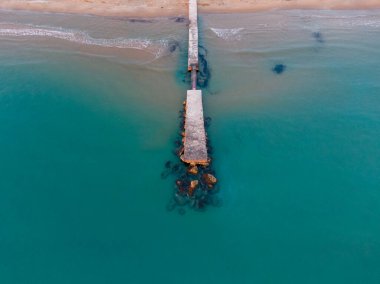 Deniz mavisi boyunca uzanan beton bir iskelenin havadan görünüşü kumlu bir kumsalla buluşuyor..
