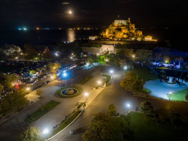 Night view of a coastal fortress illuminated, with a moonlit sea and a plaza below. clipart