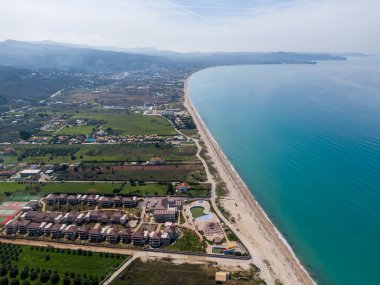 Sahil tatil köyü hava manzarası: turkuaz deniz, kumlu sahil ve konut binaları.