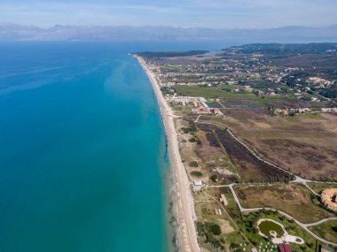 Bakir bir sahil şeridinin havadan görünüşü, turkuaz suların kumlu kıyılarla buluşması. Yeşillik ve uzak dağlar manzaralı bir manzara yaratır..