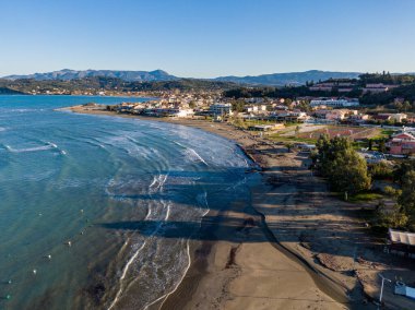 Sahil kenti sahil manzarası. Açık mavi sular kumlu sahille buluşur, arkada binalar ve dağlar..