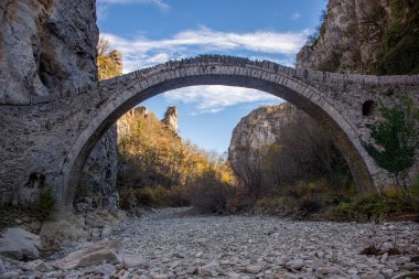 Kuru bir nehir yatağını kaplayan antik taş kemer köprüsü, yüksek uçurumlar ve sonbahar yaprakları arasına kurulmuş..