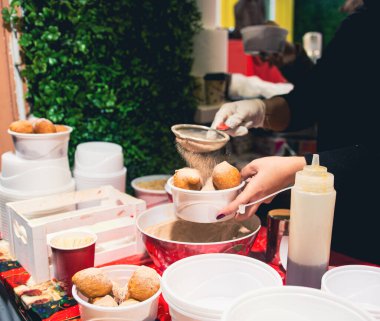 Vendor preparing delicious fried dough treats, dusting them with powdered sugar at a market.