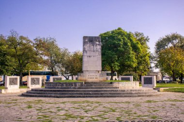Yeşillik ve açık mavi gökyüzünün altındaki ağaçlarla çevrili bir park ortamındaki taş anıt..