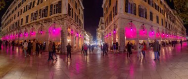 Pembe ışıklarla aydınlatılmış canlı bir şehir caddesinin gece manzarası. İnsanlar tarihi mimaride geziniyorlar..