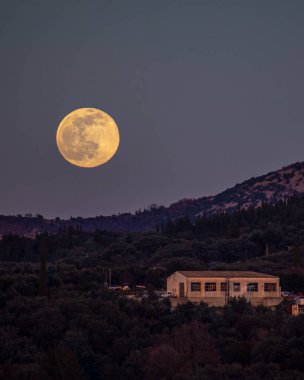 Large full moon rises over a building nestled in a hillside at twilight. clipart