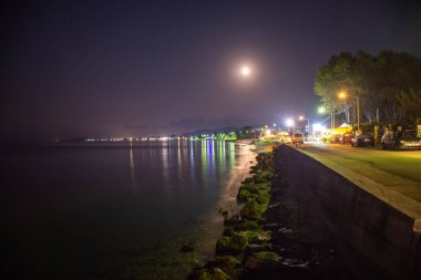 Gece sahnesi: mehtaplı kıyı kasabası, sakin deniz yansıtan ışıklar.