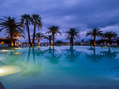 Bir tatil köyü havuzunda sakin bir akşam, suya yansıyan palmiye ağaçları....