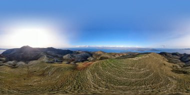 Parlak bir gökyüzünün altında kayan tepelerin ve uzak denizin panoramik görüntüsü.