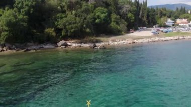 Canlı, mavi yüzen bir rıhtım, sakin, turkuaz bir suya, ağaçlarla kaplı bir sahilin arka planına ve uzaktaki küçük bir köye uzanıyor. Temiz su, denizin dibini ortaya çıkarır., 