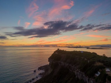 Kıyı kayalıkları ve sakin denizin üzerinde gün batımı. Dramatik gökyüzü renkleri ufku boyar.