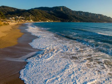 Kumlu bir sahilde dalgalar, arka planda dağlar..