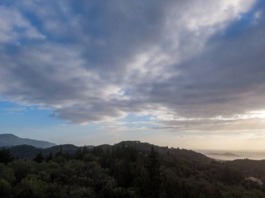 Huzurlu manzara: parçalı bulutlu bir gökyüzünün altında yuvarlanan tepeler, sakin bir denize bakan.