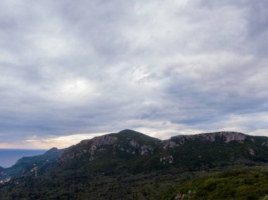 Bulutlu bir gökyüzünün altındaki dağ sıraları, kıyı manzarası.