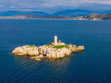 Denizde kayalık bir adada deniz feneri, arka planda dağlar.