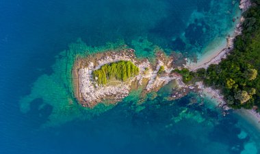 Kristal berraklığında turkuaz suyla çevrili yemyeşil bitki örtüsü olan küçük bir adanın havadan görünüşü..