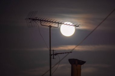 Gece gökyüzünde parlak bir dolunaya karşı siluetli anten..