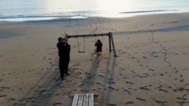 Bir kadın kumlu bir sahilde salıncakta oturuyor ve arka planda hafif dalgalar var. Biri onu filme alıyor..