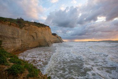 Kıyı kayalıkları bulutlu bir günbatımında dramatik dalgalarla karşılaşıyor.