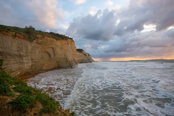 Kıyı kayalıkları bulutlu bir günbatımında dramatik dalgalarla karşılaşıyor.