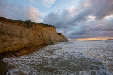 Çizgili uçurumlar bulutlu bir günbatımının altında dramatik bir okyanusla buluşur.