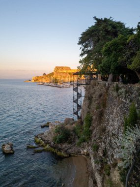 Uzakta taş bir duvar, spiral merdiven, marina ve eski mimariye sahip kıyı manzarası.