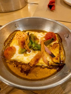 Metal bir tabakta soğan ve biberle pişirilmiş Feta peyniri. Peyniri çevreleyen kahverengi sos