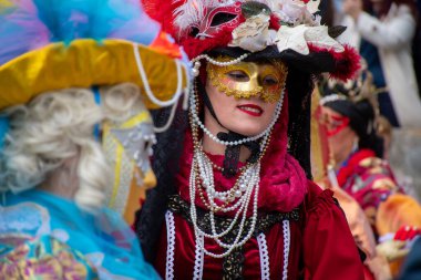 Kırmızı bir elbise, altın bir maske ve festivalde tüylü bir şapka giymiş biri..